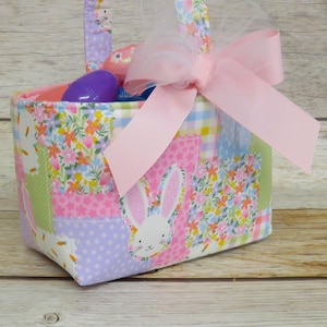 以下が含まれることがあります： パッチワークのイースターバスケット。ウサギのデザインで、カラフルな卵とピンクのリボンで飾られています。バスケットは、パステルカラーの花柄、水玉模様、ギンガムチェック柄です。ハンドルは同じパッチワーク生地で作られています。