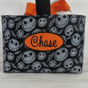 Puede incluir: Una cesta de tela negra con un estampado de caras blancas de Jack Skellington y murci&eacute;lagos. Un parche ovalado naranja con el nombre "Chase" est&aacute; cosido en la parte delantera. La cesta tiene un asa negra con un lazo naranja.