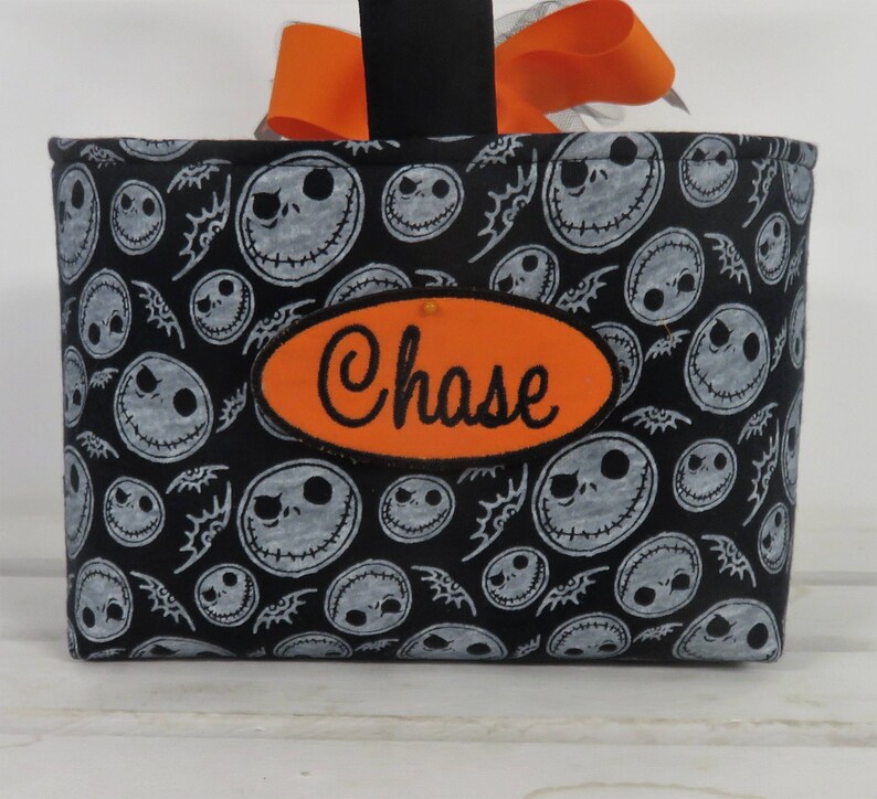 Puede incluir: Una cesta de tela negra con un estampado de caras blancas de Jack Skellington y murci&eacute;lagos. Un parche ovalado naranja con el nombre "Chase" est&aacute; cosido en la parte delantera. La cesta tiene un asa negra con un lazo naranja.