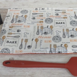 May include: A white cotton hot pad with a black and orange print of kitchen utensils and the words "Bake", "Menu", "Blend", "Eat", and "Culinaire". A red silicone spatula is also pictured.