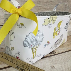 May include: A white fabric storage basket with a yellow ribbon bow. The basket features a Winnie the Pooh print with characters like Winnie the Pooh, Piglet, Eeyore, and Tigger. The basket has a gray polka dot lining and a handle.