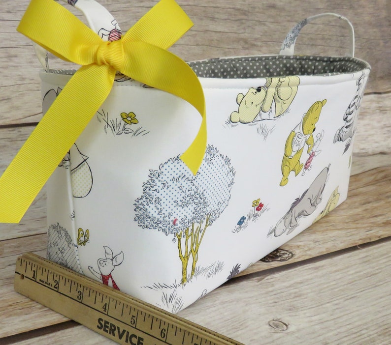 May include: A white fabric storage basket with a yellow ribbon bow. The basket features a Winnie the Pooh print with characters like Winnie the Pooh, Piglet, Eeyore, and Tigger. The basket has a gray polka dot lining and a handle.