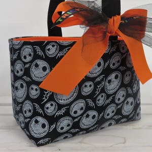 Puede incluir: Un bolso tote con tem&aacute;tica de Halloween con un fondo negro y un patr&oacute;n repetido de caras de Jack Skellington en blanco. El bolso tiene un interior naranja y est&aacute; adornado con una cinta naranja y un lazo de tul negro. La palabra "TRICK" es visible en la cinta.