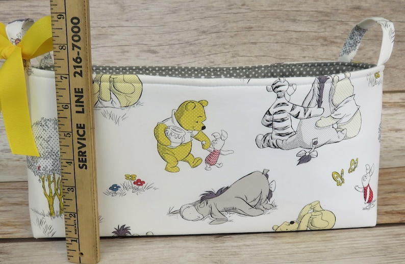 May include: White fabric storage bin with a yellow ribbon handle. The bin features a Winnie the Pooh theme with illustrations of Winnie the Pooh, Piglet, Eeyore, Tigger, and other characters from the Disney cartoon.