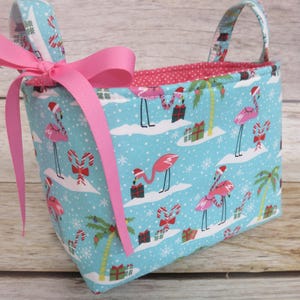 May include: A rectangular fabric basket with a festive flamingo print in a winter scene. The basket features a light blue background with pink flamingos wearing Santa hats, candy canes, and wrapped gifts. It has pink handles and a pink bow.