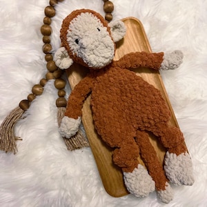 May include: A handmade, plush monkey toy in shades of brown and cream. The monkey is lying on a wooden tray, with a beaded garland and tassel in the background. The toy has a textured, fuzzy appearance.