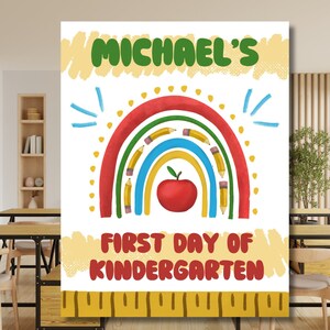 Puede incluir: Un colorido letrero de jardín de infancia con el nombre "Michael's" en verde, sobre un diseño de arcoíris con lápices y una manzana roja. El texto "First Day of Kindergarten" está en rojo. Un diseño de regla amarilla en la parte inferior.