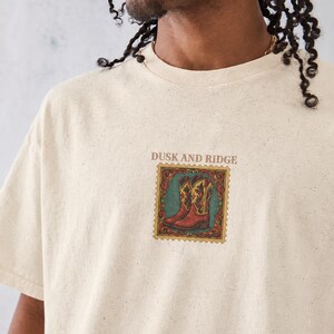 May include: A cream-colored short-sleeve t-shirt with a speckled texture. The center of the chest features a graphic of two cowboy boots inside a decorative postage stamp frame, with the text DUSK AND RIDGE printed above the graphic.