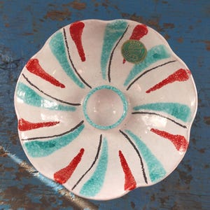 May include: A white ceramic serving dish with a scalloped edge. The dish features a central dip bowl and is decorated with red, teal, and black abstract designs. A green and gold sticker is visible on the dish.