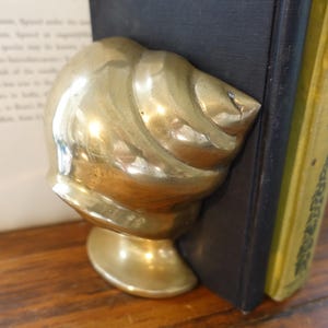 May include: A brass bookend in the shape of a seashell. The bookend is positioned next to a black book and a book with a yellow cover. The seashell design has detailed ridges and a smooth, polished finish.