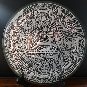 May include: A round, dark gray decorative plate with intricate silver-colored etched designs. The plate features a central image of a deer and various human figures, surrounded by geometric patterns and symbols. The plate is displayed on a black stand.