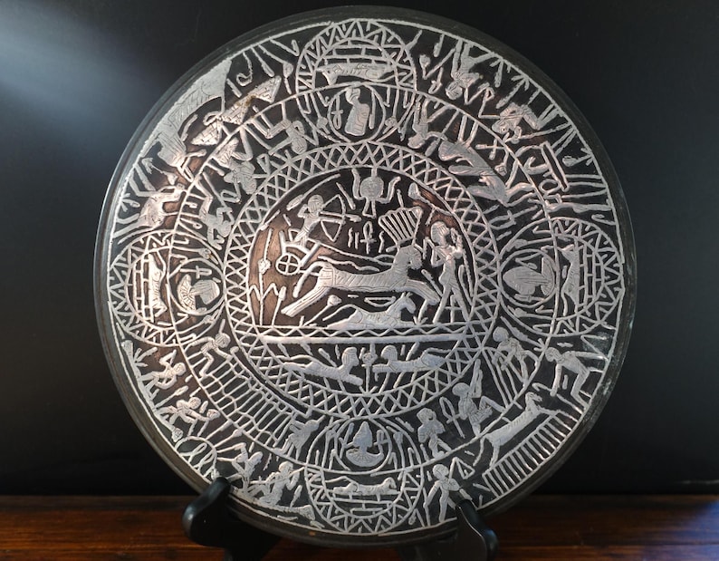 May include: A round, dark gray decorative plate with intricate silver-colored etched designs. The plate features a central image of a deer and various human figures, surrounded by geometric patterns and symbols. The plate is displayed on a black stand.