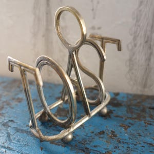 May include: A silver-colored metal toast rack. The rack is shaped to spell out the word "TOAST" in a stylized font. The rack has a rectangular base with small ball feet and a handle. The background is a blue surface.