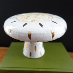 May include: A white ceramic mushroom-shaped decorative object with gold leaf accents. The object has a wide, flat top with a recessed center and a rounded stem. The gold leaf accents are in a teardrop shape. The object is sitting on a green surface.
