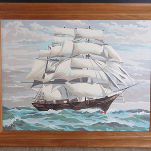 May include: A framed painting of a large sailing ship with white sails navigating through choppy, teal and white waves under a pale, cloudy sky. The artwork is enclosed in a simple, light brown wooden frame with a beveled edge design.