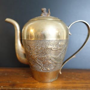 May include: A brass teapot featuring a smooth upper section and a textured lower body with an embossed dragon motif. The lid is topped with a small animal figurine, and the piece includes a curved handle and a long, slender spout.