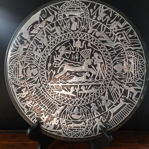 May include: A round, dark-colored decorative plate with intricate silver-colored etched designs. The plate features a central scene with figures and animals, surrounded by concentric circles of additional figures and patterns. The plate is displayed on a black stand.