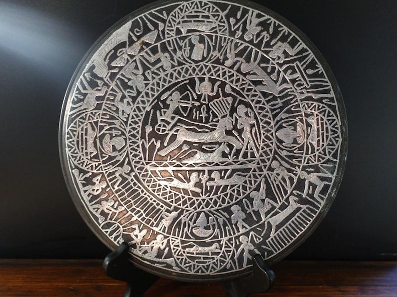 May include: A round, dark-colored decorative plate with intricate silver-colored etched designs. The plate features a central scene with figures and animals, surrounded by concentric circles of additional figures and patterns. The plate is displayed on a black stand.