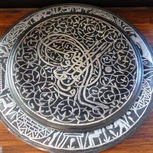 May include: A black decorative plate with intricate silver-colored patterns. The plate features a central design with swirling lines and floral motifs, surrounded by a detailed border. The plate is likely made of metal and is displayed on a wooden surface.