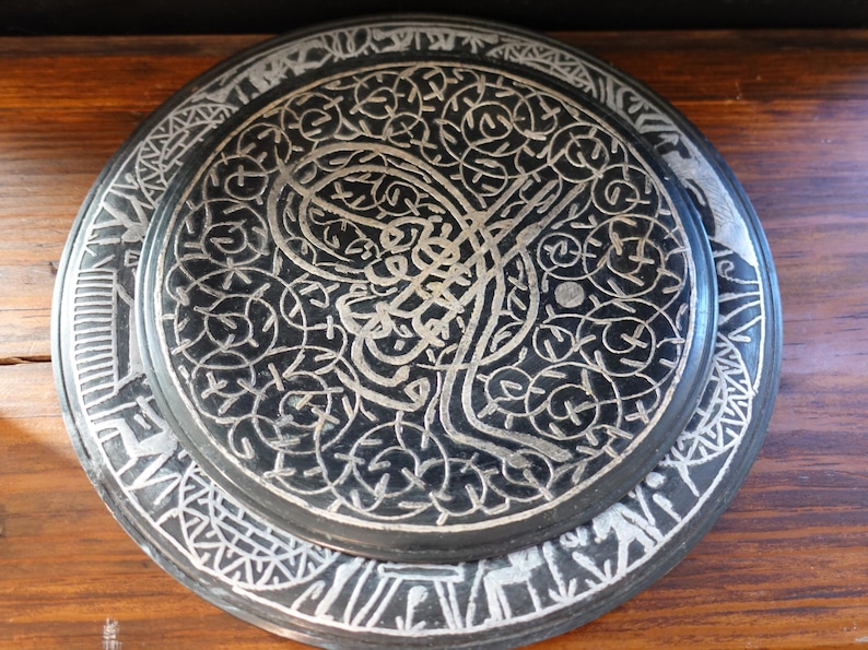 May include: A black decorative plate with intricate silver-colored patterns. The plate features a central design with swirling lines and floral motifs, surrounded by a detailed border. The plate is likely made of metal and is displayed on a wooden surface.