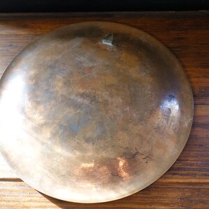 May include: A vintage, round, bronze-colored metal bowl with a slightly concave shape. The bowl has a tarnished, aged appearance with variations in color, including brown and hints of blue. A small, triangular decorative element is visible.