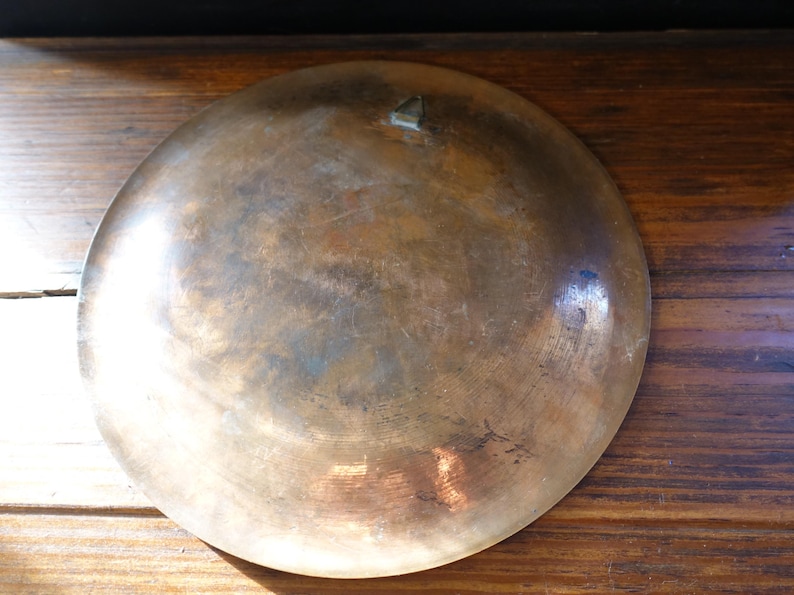 May include: A vintage, round, bronze-colored metal bowl with a slightly concave shape. The bowl has a tarnished, aged appearance with variations in color, including brown and hints of blue. A small, triangular decorative element is visible.