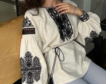 May include: A white linen blouse with black embroidery. The blouse has a round neckline, long sleeves, and a tie at the waist. The embroidery is intricate and features floral and geometric patterns.