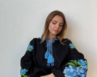 Op de afbeelding: Een zwarte geborduurde blouse met blauwe bloemenpatronen en blauwe kwastjes. De blouse heeft lange mouwen en een ronde hals.