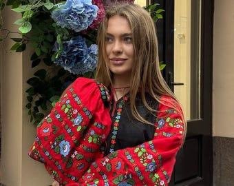 May include: A black blouse with red sleeves featuring floral embroidery in blue, green, and white. The blouse has a round neckline and is worn with blue jeans. The background includes colorful flowers.