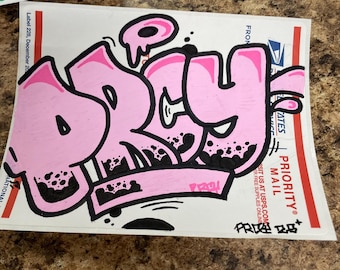 Puede incluir: Una obra de arte de estilo graffiti rosa sobre un fondo blanco. La palabra "DRY" está escrita en letras de burbujas con contornos negros y sombreado moteado negro. La obra de arte está en una etiqueta USPS Priority Mail.