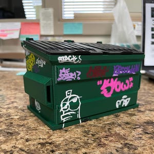 Peut inclure: Une benne à ordures miniature vert foncé avec un couvercle noir articulé et divers marquages de style graffiti en peinture blanche, rose et violette. L'extérieur présente des illustrations et du texte dessinés à la main, dont le mot Brooklyn en violet.