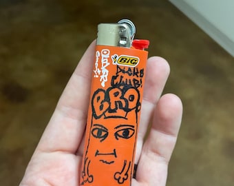 Könnte beinhalten: Orangener Feuerzeug mit einer darauf gezeichneten schwarzen Comicfigur. Die Figur hat einen großen Kopf mit Augen, einem Mund und einer Krone. Auf dem Feuerzeug steht der Text "Old Skool" und "Dicks Club!" Das Feuerzeug ist von der Marke BIC.