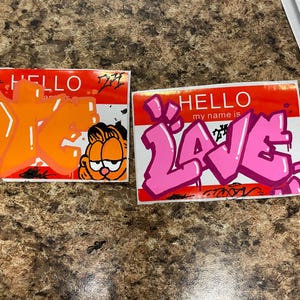 Peut inclure: Deux autocollants de type étiquette nominative avec du graffiti. L'un présente du texte orange et un dessin animé de Garfield. L'autre a le mot "LOVE" en rose. Les deux ont un fond rouge et blanc avec le mot "HELLO".