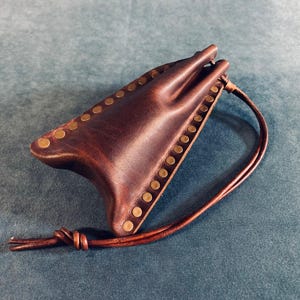 May include: A brown leather pouch with brass rivets along the edges. It features a drawstring closure and a knotted leather cord. The leather is a rich, dark colour with a slightly textured appearance. The pouch is designed to hold small items.