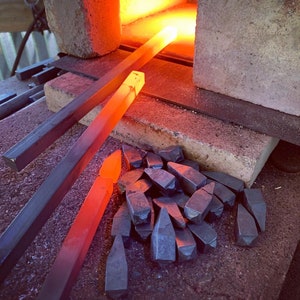 Op de afbeelding: Een close-up van een smederij met twee metalen staven die in het vuur worden verhit. Op de voorgrond ligt een stapel metalen stukken.