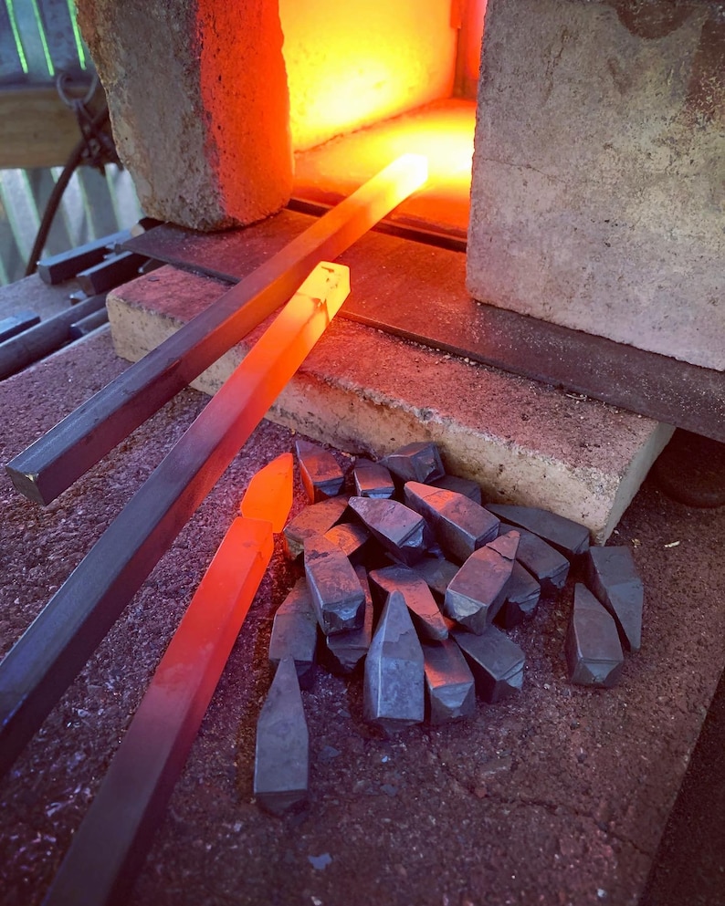 Op de afbeelding: Een close-up van een smederij met twee metalen staven die in het vuur worden verhit. Op de voorgrond ligt een stapel metalen stukken.