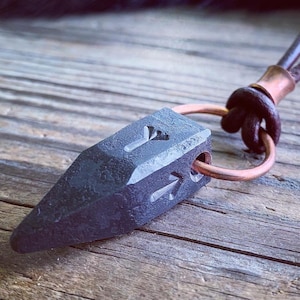 Op de afbeelding: Een zwarte metalen hanger in de vorm van een puntige pijlpunt met een koperen ring eraan bevestigd. De hanger heeft een getextureerd oppervlak en is gegraveerd met een symbool.