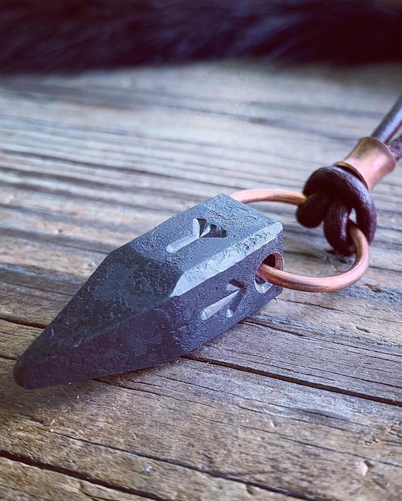 Op de afbeelding: Een zwarte metalen hanger in de vorm van een puntige pijlpunt met een koperen ring eraan bevestigd. De hanger heeft een getextureerd oppervlak en is gegraveerd met een symbool.