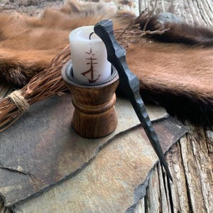 May include: A black metal wand with a pointed tip, resting on a slate surface next to a wooden candle holder with a white candle. The candle has a symbol etched into it.