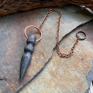 Könnte beinhalten: Ein schwarzes Metallpendel mit einer spitzen Spitze und einer Kupferkette. Die Kette hat oben einen Ring und unten einen kleineren Ring.
