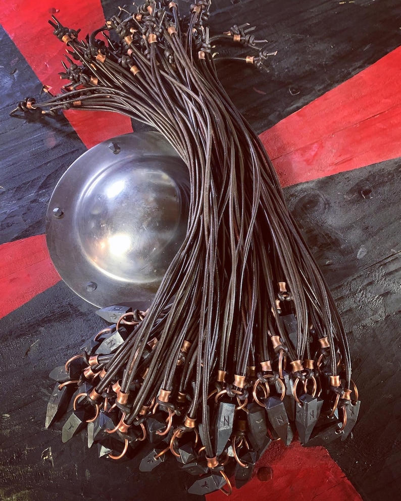 Op de afbeelding: Een bundel zwarte leren koorden met koperen accenten en zwarte metalen pijlpunt hangers. De koorden zijn aan de bovenkant aan elkaar gebonden en de pijlpunten hangen naar beneden.