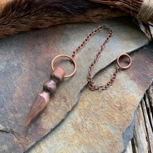 May include: A copper pendulum with a pointed tip and a chain with a ring at the end. The pendulum is resting on a textured, gray surface.