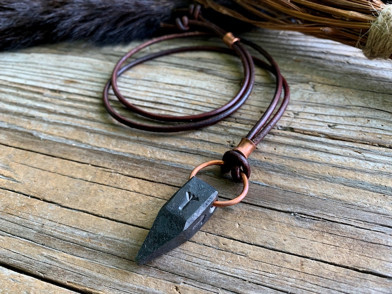 Op de afbeelding: Een ketting van zwart lederen koord met een koperen sluiting en een zwarte stenen hanger met een gegraveerde rune.