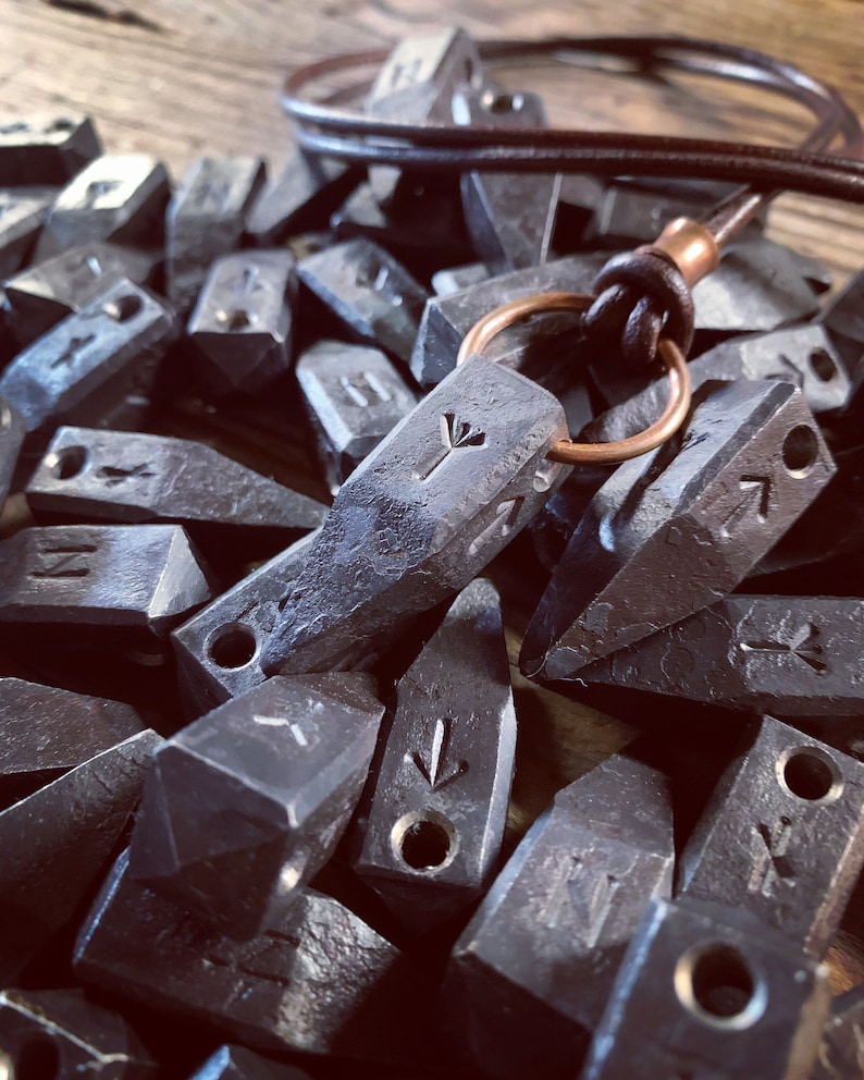 Op de afbeelding: Een close-up van een ketting met een hanger van metaal. De hanger is gevormd als een klein, puntig mes en heeft een gegraveerd symbool. De ketting is gemaakt van bruin lederen koord en heeft een koperen ring aan de bovenkant. De hanger wordt omringd door een stapel vergelijkbare metalen hangers, elk met een ander gegraveerd symbool.