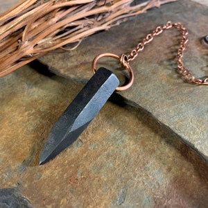 Könnte beinhalten: Ein schwarzes Metallpendel mit einer spitzen Spitze, befestigt an einer Kupferkette mit einem Ring am Ende. Das Pendel liegt auf einer grauen Steinoberfläche.