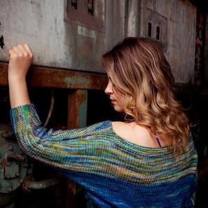 Peut inclure: Une femme aux longs cheveux bruns ondulés porte un pull rayé bleu, vert et jaune. Elle se tient devant un mur métallique rouillé avec un panneau de commutateurs.
