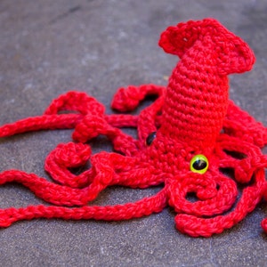 May include: A crocheted red squid with green eyes. The squid has multiple tentacles and is lying on a gray surface.