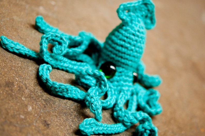 May include: A crocheted turquoise squid with a green eye. The squid has eight tentacles and is lying on a brown surface.