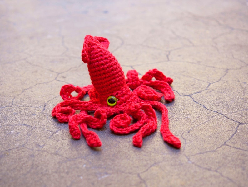 May include: A crocheted red squid with a single green eye. The squid has many tentacles and is lying on a cracked, gray surface.