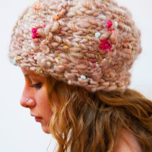 May include: A woman with curly brown hair is wearing a beige knitted beanie with colorful embellishments. The beanie has a slouchy fit and is decorated with pink, blue, and gold accents.